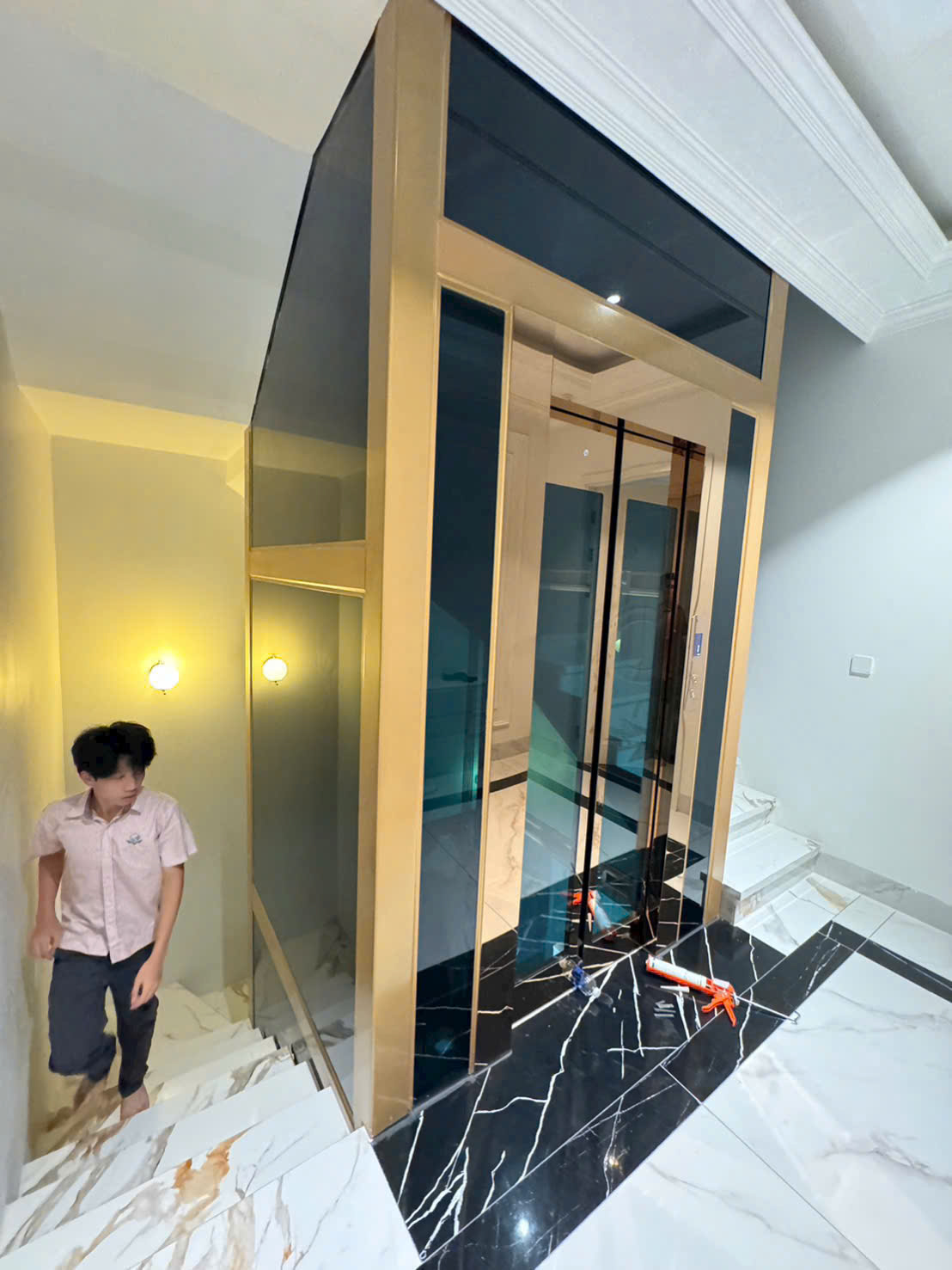 Thang máy gia đình khung kính viền vàng lắp đặt cạnh cầu thang trên nền đá hoa cương